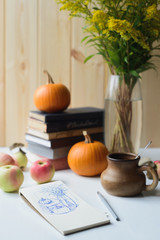 Obraz na płótnie Canvas sketch in notebook of autumn still life with books stacked, orange pumpkins, apple, clay drinking cup with spoon, solidago bouquet, side view of vertical stock photo image