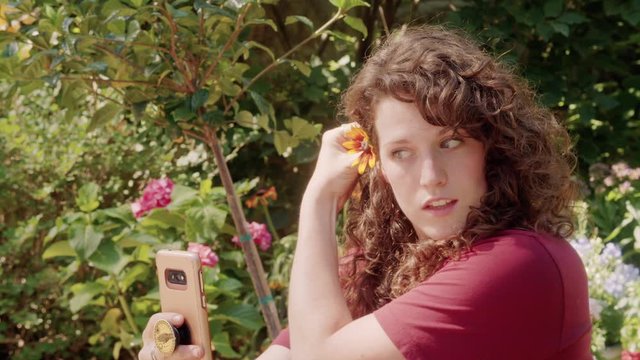 A Young woman with flower in hair takes selfies with her phone in a garden