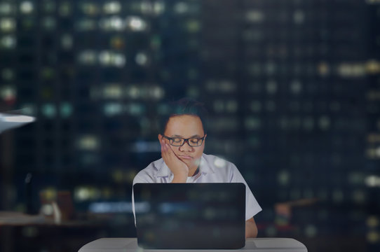 Young male student in school uniform wearing eye glasses falling asleep while doing his project and research in front of his laptop. Studying at night inside office room. 