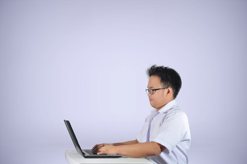 Side view shot of a young male student wearing white polo school uniform working and studying using his laptop on his desk in white isolated background. 