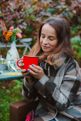 young woman in a brown coat sits in a chair at a table with a red mug draped in a plaid outdoors...
