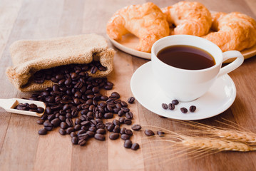 Top view white hot coffee cup and beans on wooden table with croissant bread background.Black coffee menu for breakfast in the coffee shop.Selective focus.