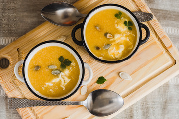 Pumpkin puree soup decorated with pumpkin seeds and cream in two bowls on a wooden tray and spoons top view