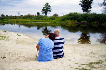 Happy senior couple sitting by the river at sand, calm evening