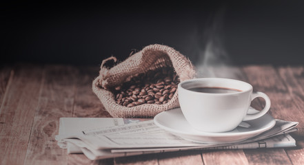 Coffee cup with smoke rising,roasted coffee beans and newspaper on wood table with dark background