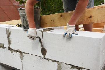 Bricklayer builder laying autoclaved aerated concrete blocks for house wall near unfinished roof wooden beams.