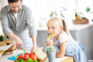 Blonde girl eating yummy pepper while father cooking lunch