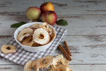 Dried apples chips in bowl with fresh red apples and cinnamon sticks on table.
