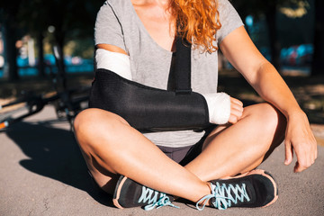A young red-haired woman sits in sportswear on a bicycle path with a broken arm bandaged in a cast...