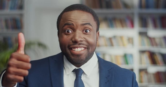 Close up of the cheerful and happy African American young businessman in suit and tie smiling to the camera while giving his thumb up in the library. Portrait.