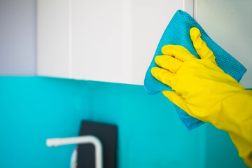 A female hand in yellow rubber gloves wipes a modern white kitchen with a microfiber cloth.