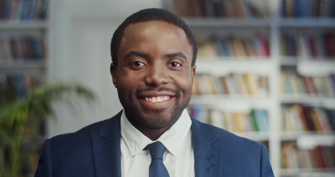 Portrait shot of the attractive and cheerful young African American businessman looking at the side, then turning his face to the camera and laughing in the library. Close up.