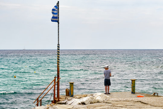 Young boy in a sailor t-shirt with a fishing rod fishing on the background of the sea on the dock of Scala Potamias, Tassos, Greece, in summer, alone