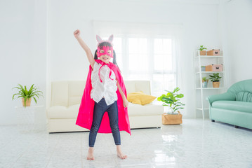 Happy little asian girl wearing pink superhero clothing standing on living room floor and posing...