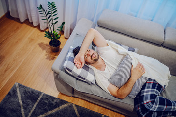 Top view of very sick caucasian man in pajamas and covered with blanket lying on sofa in living room, holding pillow and having stomachache.