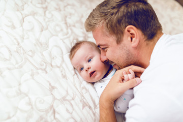 Close up of beautiful young caucasian dad cuddling with his six months old son. Baby lying in bed and hugging father.