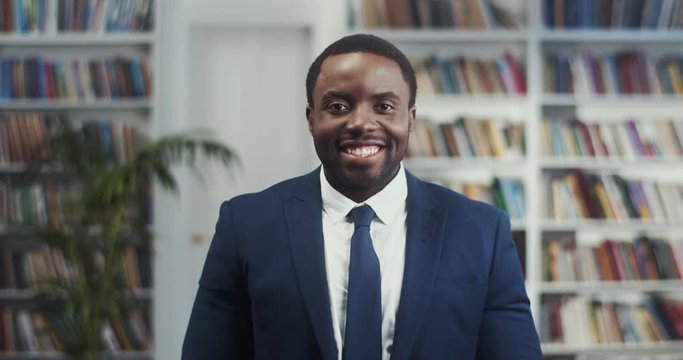 Portrait shot of the young African American handsome businessman in suit and tie standing in the library and smiling cheerfully.