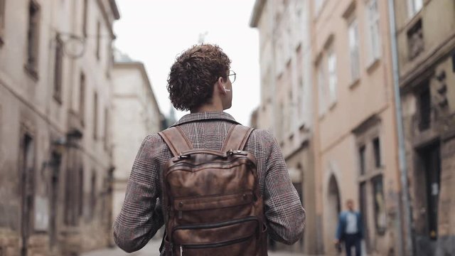 Back View of Young Tourist Guy with Brown Backpack and Earphones Wearing Checked Jacket Walking through Old City Street, Looking Around.