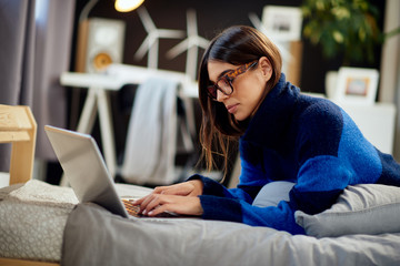 Fototapeta na wymiar Attractive caucasian brunette in blue and black sweater lying on stomach in bed and typing on laptop.