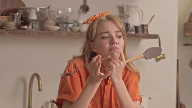 Pensive pretty young blonde woman holding kitchen utensils and thinking about something while cooking at the kitchen
