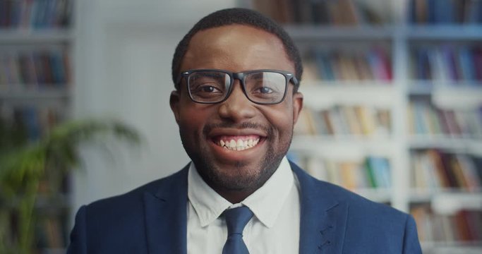 Portrait shot of the young happy and handsome African American businessman in glasses, suit and tie looking straight to the camera and laughing cheerfully in the library. Close up.