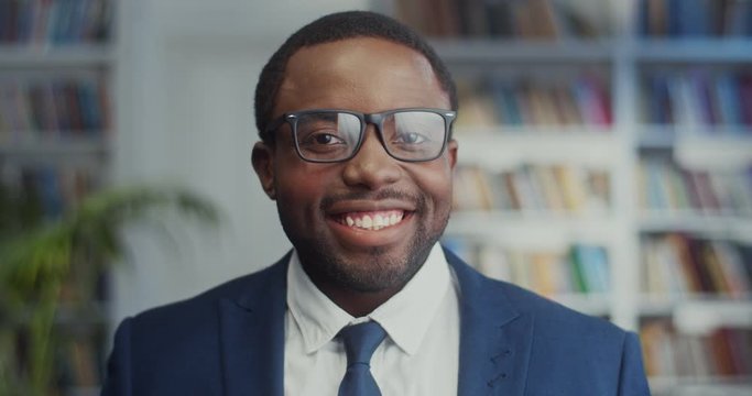 Portrait of the young attractive African American man in business style: suit, tie and glasses turning face to the camera and smiling joyfully in the library. Close up.