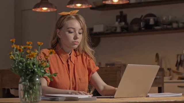 Sleeping cute young blonde woman waking up and continuing working with laptop while sitting at the kitchen