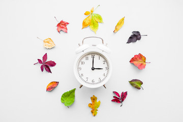 Colorful different autumn leaves around white alarm clock on light gray background. Time change...