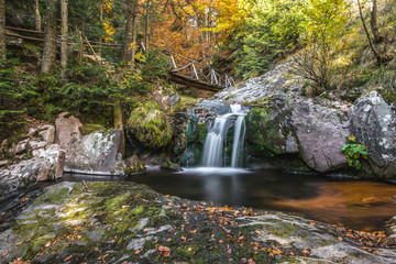 Fototapeta na wymiar Small creek waterfall in a beautiful deciduous autumn forest. Bright autumn leaves on stones covered with moss by the river. Beautiful autumn landscape with wooden bridge, Long exposure.