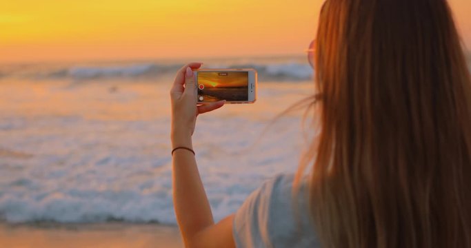 Young female felling fun and free on beach at sunset. Carefree girl on tropical beach takes photos of nature on a smartphone at golden sunrise. Happy tourist woman on summer vacation. 5g Internet.