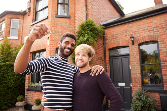 Portrait Of Excited Gay Male Couple Standing Outside New Home Holding Keys On Moving Day Together