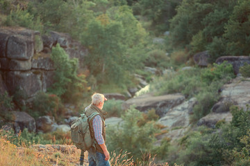 Eldery gray-haired tourist with backpack is walking on rocks on the background of canyon