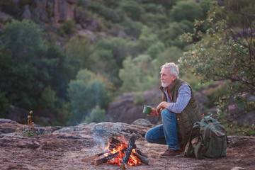 Eldery tourist with gray hair and beard sits on the edge of a canyon near a burning fire with a mug in his hands, against the backdrop of the sun, mountains and trees