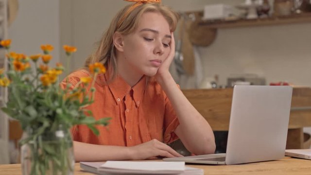 Exhausted young blonde woman becoming tired and falling asleep while working with laptop sitting at the kitchen