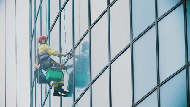 A man worker hanging on ropes and wipes the exterior windows of a business skyscraper - industrial alpinism - overcast weather