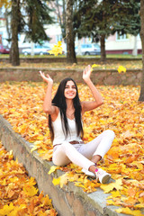 Beautiful girl in autumn park sitting on a concrete parapet catches falling maple leaves with her hands a smile on her face, in white jeans and a blouse against a background of trees  yellow foliage