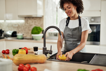 Charming mixed race woman in apron standing in kitchen ad washing yellow paprika in sink. On kitchen counter are different sorts of vegetables.