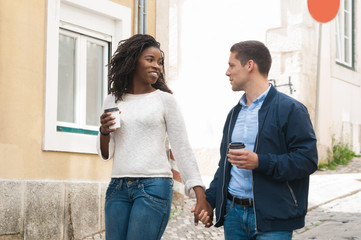 Happy multiethnic couple walking outside. Young man and woman going down old street, holding takeaway coffee cup and talking. Walking outside concept