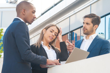 Multiethnic business partners watching and discussing presentation outside. Business men and woman standing near office building, using laptop, looking at screen, talking. Colleagues meeting concept