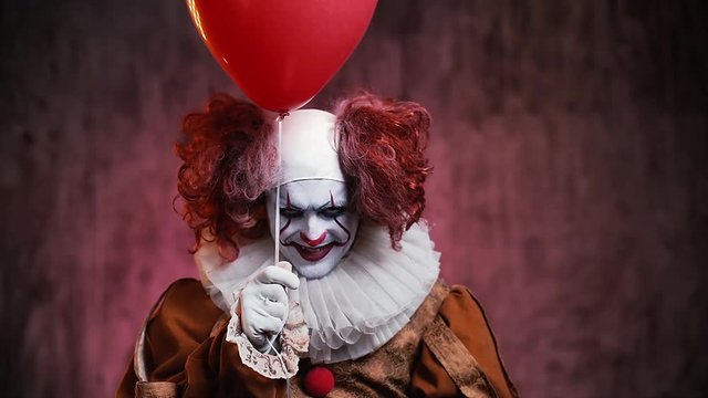 Creepy clown with colorful make-up is looking into the camera, holds a red ball in his hand and welcomes the viewer. Horrifying clown is standing in a dark room smiling.