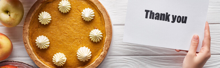 cropped view of woman holding thank you card near pumpkin pie on wooden white table with apples, panoramic shot