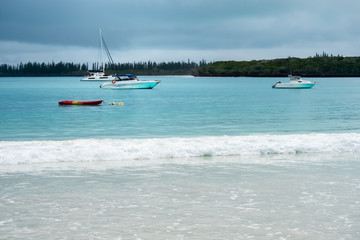 Fototapeta na wymiar Boats on the water and waves crashing on the beach at Kuto Bay with pine forests at the horizon in the background on a beautiful day at Isle of Pines in New Caledonia, South Pacific Ocean.