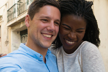 Happy excited mix raced couple having fun outside. Closeup of young man and woman standing in old town street, looking away, smiling, laughing. Dating couple concept