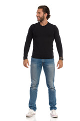 Handsome Man In Black Jersey, Jeans And Sneakers Is Standing Relaxed, Looking Away And Smiling. Front View.