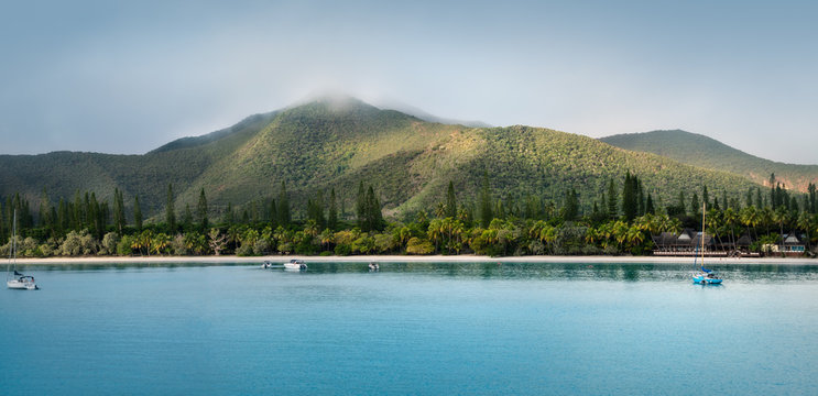 Kuto Bay Panorama with tropical vegetation, white sandy beach with boats on the water and Pic N'Ga Mountain top engulfed in clouds and sun-lit mountain ridges on Isle of Pines, New Caledonia.