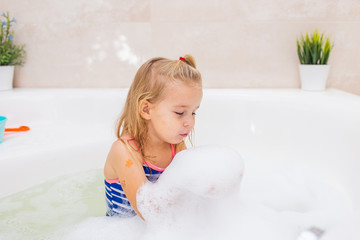 Little blonde smiling girl taking bubble bath in beautiful bathroom.Kids hygiene. Shampoo, hair treatment and soap for children. Kid bathing