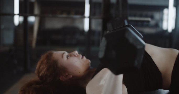 Close-up young beautiful Caucasian red haired athletic woman lifting dumbbells lying on bench in large professional gym.
