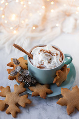 Hot winter drink: chocolate with whipped cream in blue mug. Christmas time. Cozy home atmosphere, white background. Homemade gingerbread cookies, cones, candle, lights as decor. Holiday festive mood