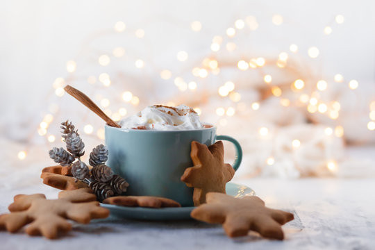 Hot winter drink: chocolate with whipped cream in blue mug. Christmas time. Cozy home atmosphere, white background. Homemade gingerbread cookies, cones, candle, lights as decor. Holiday festive mood