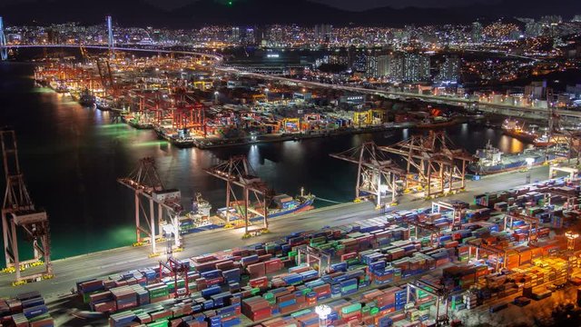 Timelapse Busan operating busy cargo port with illuminated colourful containers ready for shipment and colours reflection on water at night against city zoom in